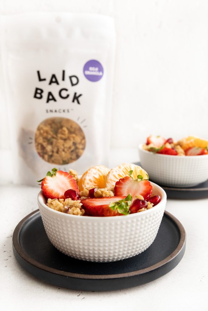 Laid Back Snacks parfait bowl with goji granola, and granola in a white bowl with strawberries and oranges and fruits.